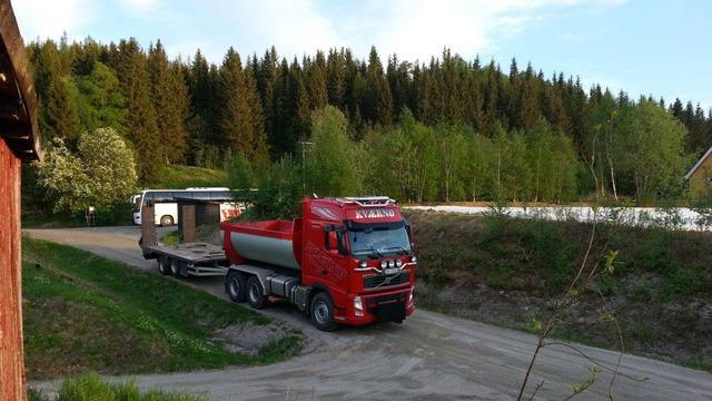 En rød lastebil med tilhenger står på en grusvei omringet av grønne trær og skog.