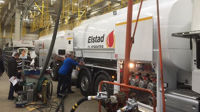 Lastebiler med tank for drivstoff fylles opp i en industriell setting med arbeidere som jobber.