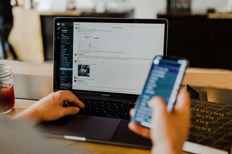 En person skriver på en laptop mens de holder en mobiltelefon i hånden, med en skjerm som viser en chat-applikasjon.