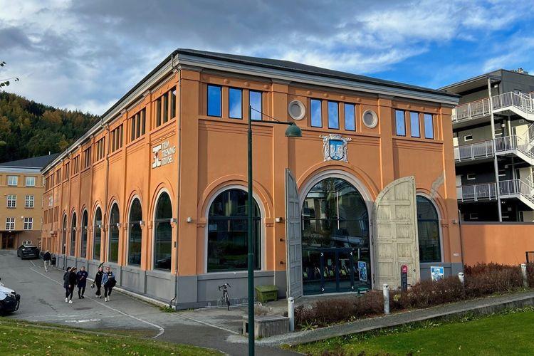 En stor, oransje bygning med buede vinduer og moderne detaljer, omgitt av grønt landskap og mennesker som går forbi.