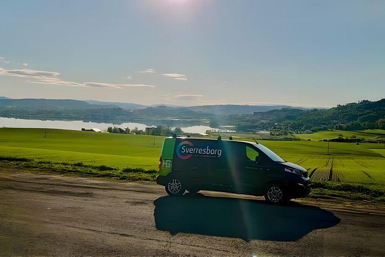 En svart varebil med logoen "Sverresborg" står langs en vei med grønt landskap og innsjø i bakgrunnen.