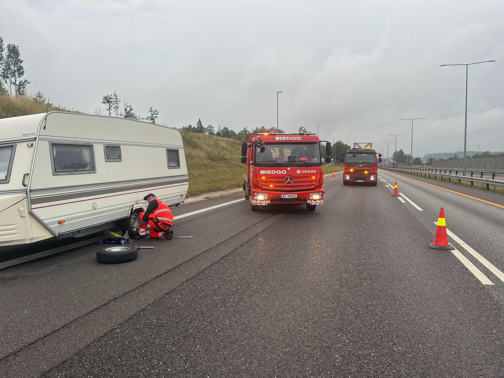 En rød brannbil og en campingvogn på veien, med en person som skifter dekk. Advarselstriangler er plassert langs veien.