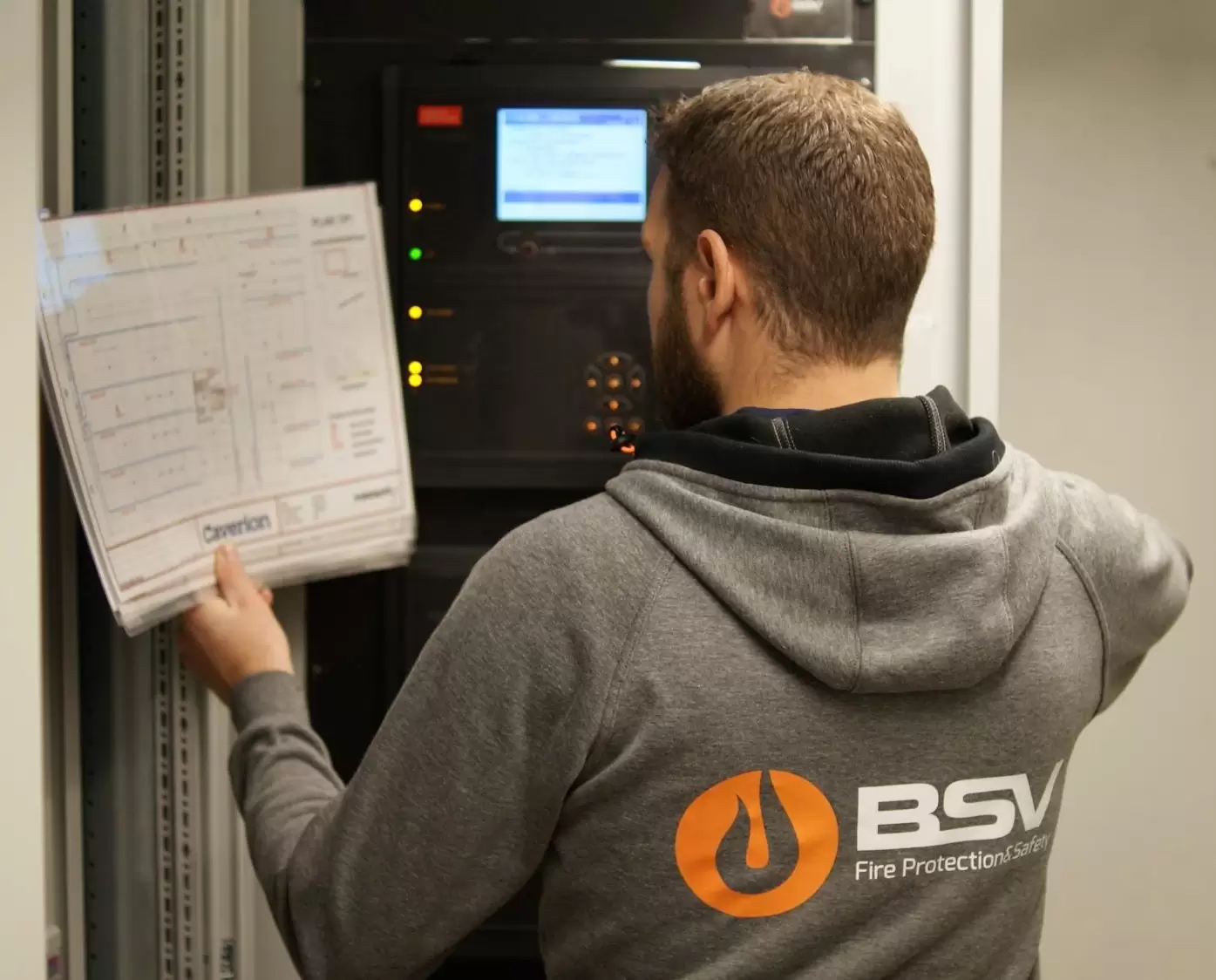 En tekniker som inspiserer brannalarmsystemet med en plan i hånden, iført en hettegenser med BSV-logo.