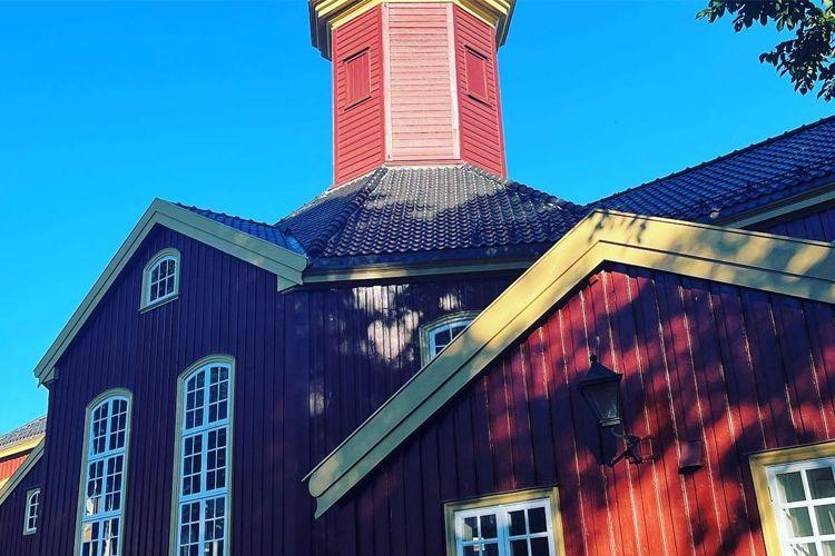 Rødt hus med gult tak og tårn, omgitt av trær og blå himmel. Arkitektoniske detaljer er synlige.