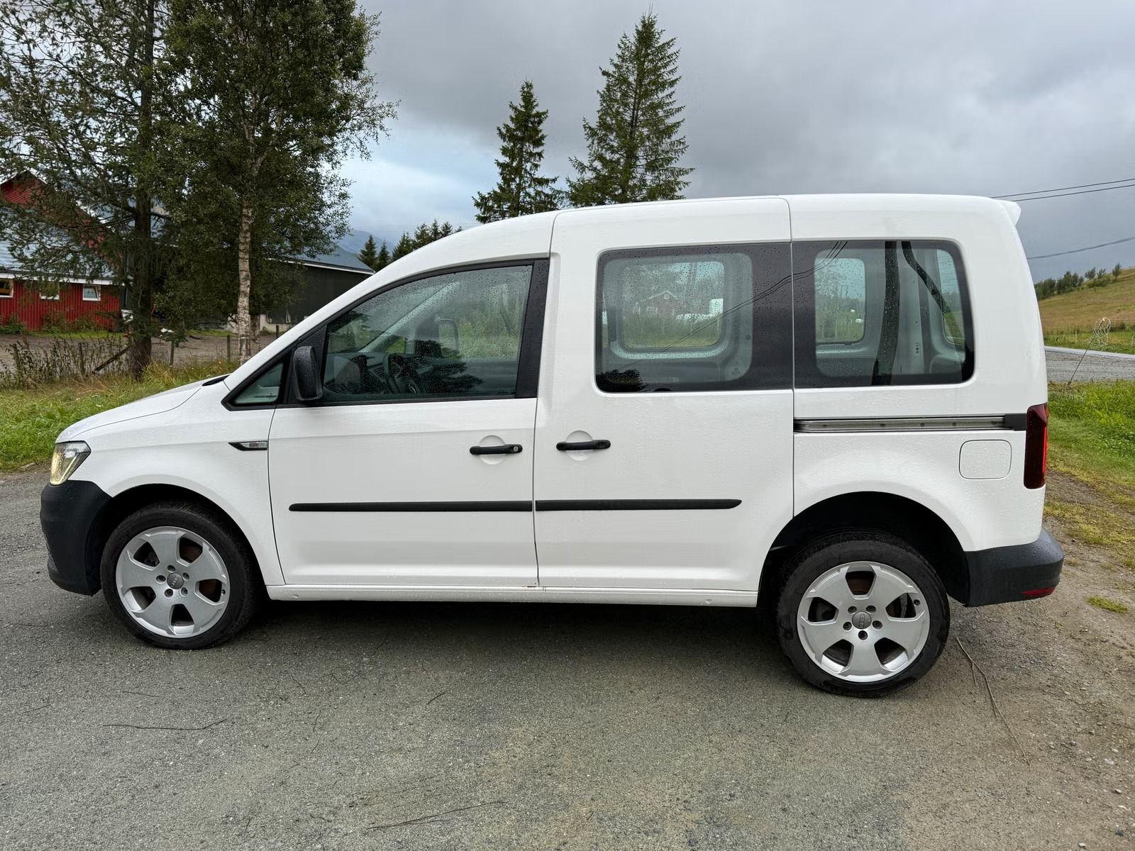 Hvit varebil parkert på en vei med trær og bygninger i bakgrunnen.