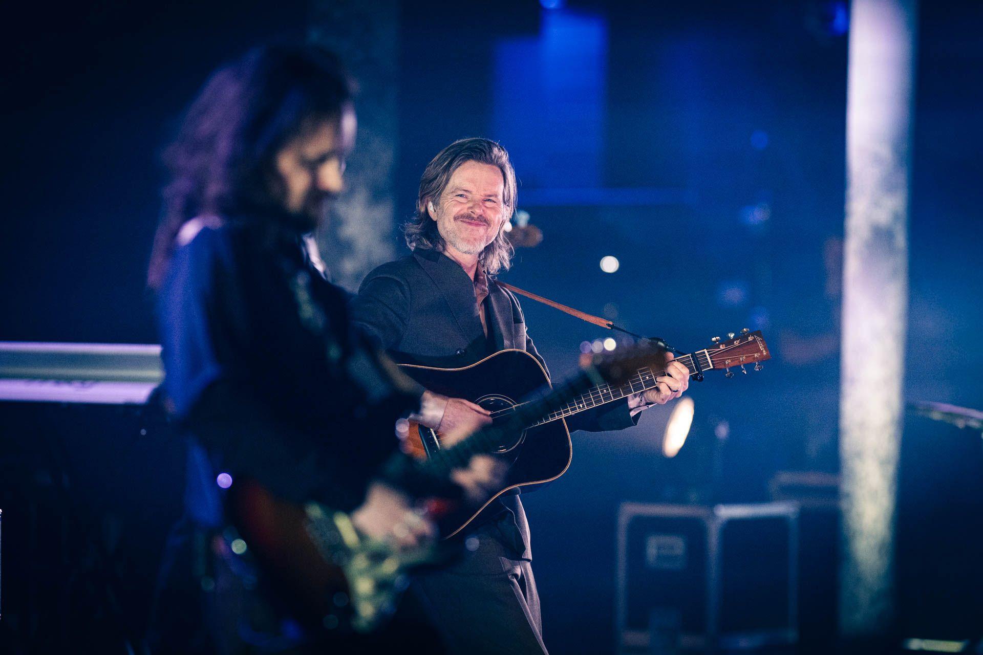 To musikere som spiller gitar på en scene med blå belysning under en konsert.