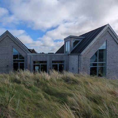 Moderne hus med grå murstein og store vinduer, omgitt av høyt gress under en blå himmel.