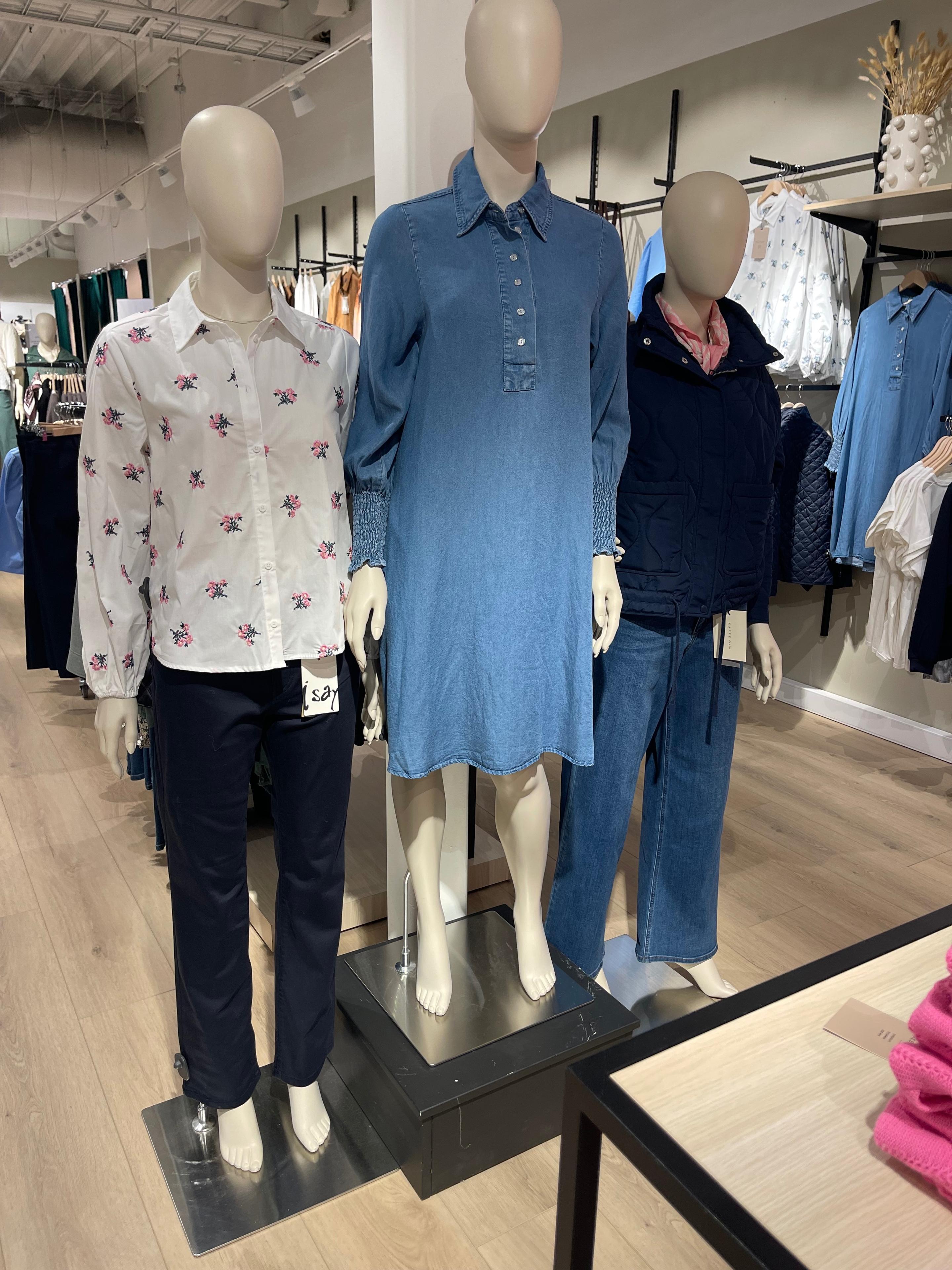 Tre mannequiner i en butikk, iført en bluse, en denimkjole og en jakke med bukser.
