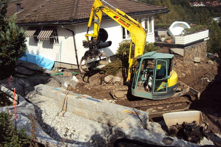 Gravemaskin i arbeid på byggeplass ved hus, med stein og jord rundt.