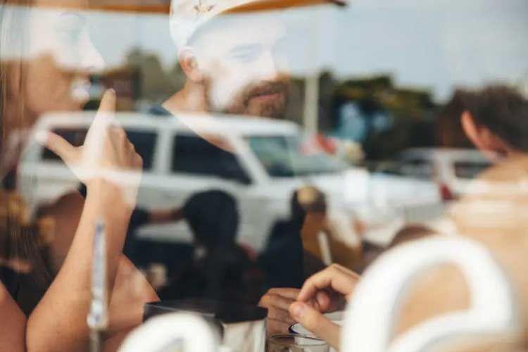 To personer som sitter ved et bord i en kafé, med refleksjoner av biler utenfor vinduet.