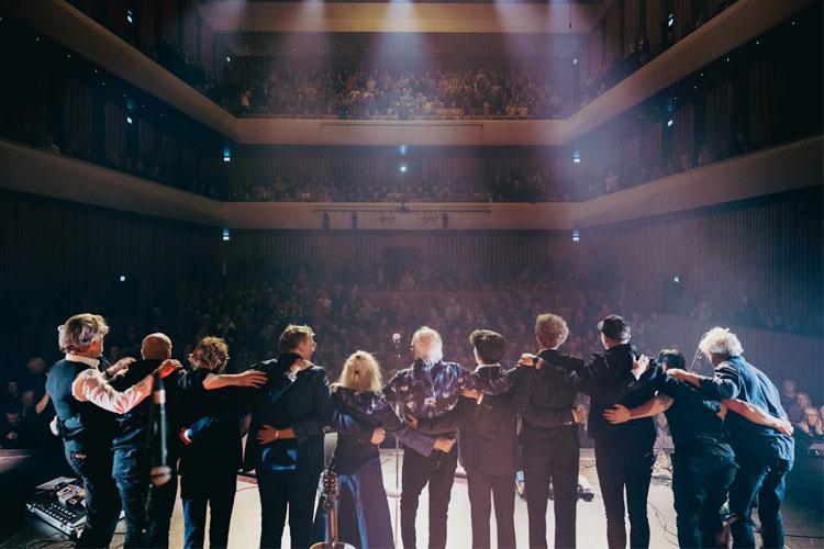 En gruppe musikere står på scenen og tar imot applaus fra publikum i en stor konsertsal.