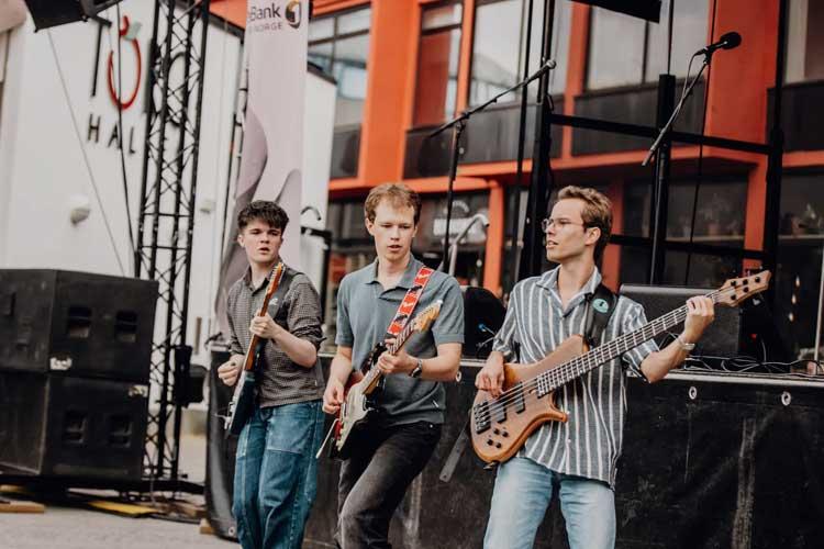 Tre unge musikere spiller gitar på en utendørskonsert med scenen i bakgrunnen.