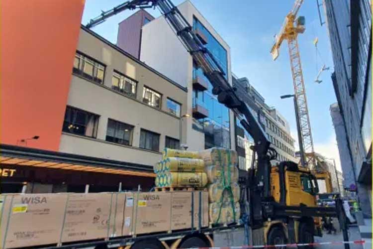 Lastebil med kran som løfter byggevarer foran en bygning i en bygate. Kranen er i bruk.