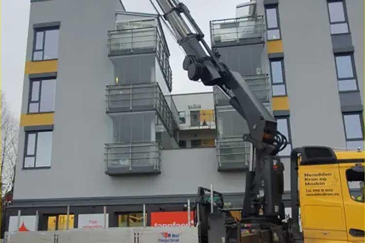 En kran løfter balkonger til en ny bygning med grå fasade og gule detaljer. Byggeplassen er avsperret.