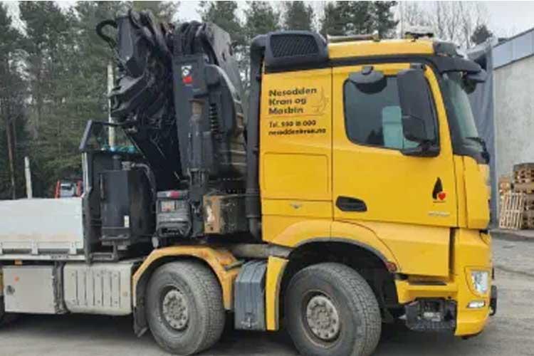 Gul lastebil med kran, parkert ved en bygning, omgitt av trær og materialer.