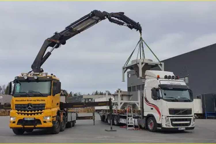 To lastebil med kran løfter en container fra en annen lastebil på en byggeplass.