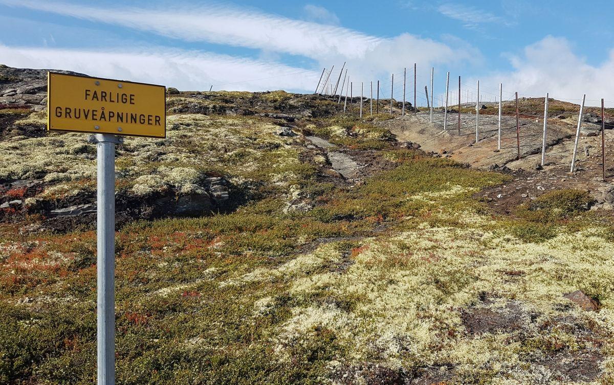 Farlig brulehninger skilt ved en steinete bakke med grønt lyng og blå himmel i bakgrunnen.
