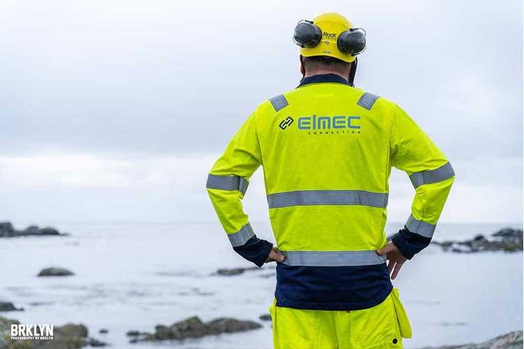 En arbeidstaker i refleksjakke står ved kysten og ser utover havet, med sikkerhetsutstyr på hodet.