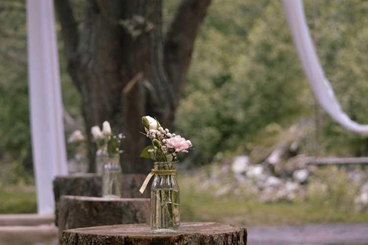 Vaser med blomster på trestammer i en utendørs bryllupssetting med hvite stoffer i bakgrunnen.