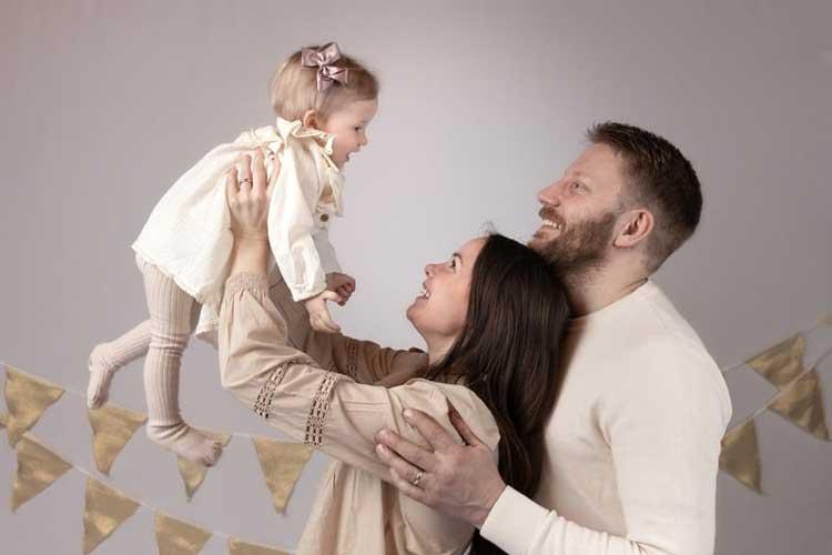 Familieportrett med foreldre som løfter en liten jente i en hvit kjole, smilende mot hverandre.