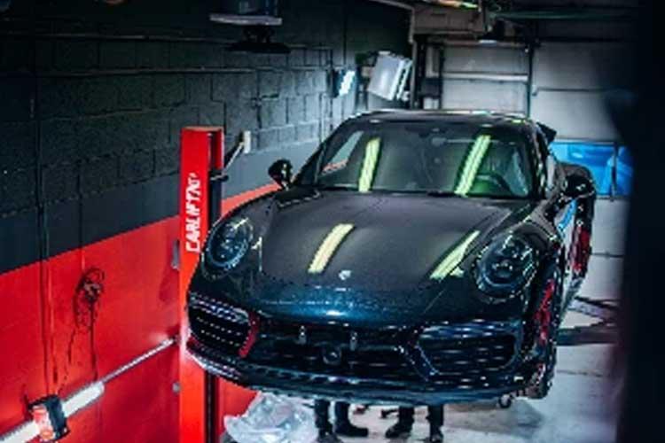 En svart Porsche 911 heves på en bilheis i et verksted med røde og svarte vegger.