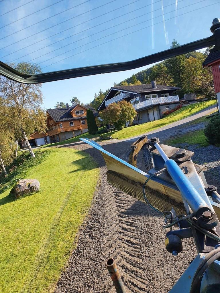 Utsikt fra en traktor som pløyer en hagevei med hus i bakgrunnen og grønt gress på siden.