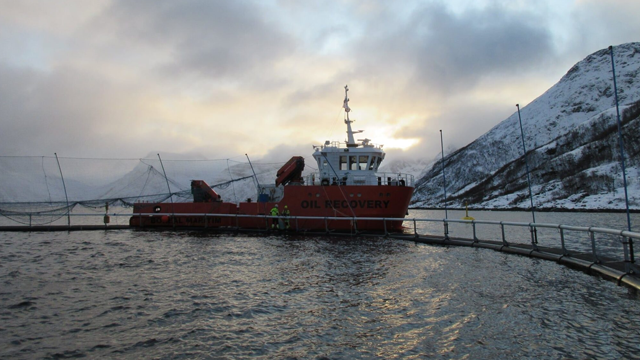 En oljeutvinningsbåt ligger ved en kai i fjorden, omringet av snødekte fjell og en dramatisk himmel.