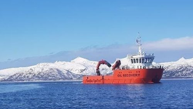 En oransje oljeutvinningsbåt seiler på havet med snødekte fjell i bakgrunnen.