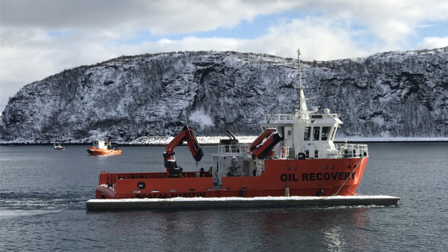 En oransje oljeoppsamlingsbåt seiler på vannet med snødekte fjell i bakgrunnen.