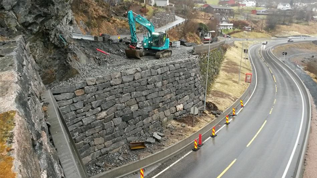 Gravemaskin arbeider på en steinmur langs veien, med veiarbeidsskilt og landskap i bakgrunnen.