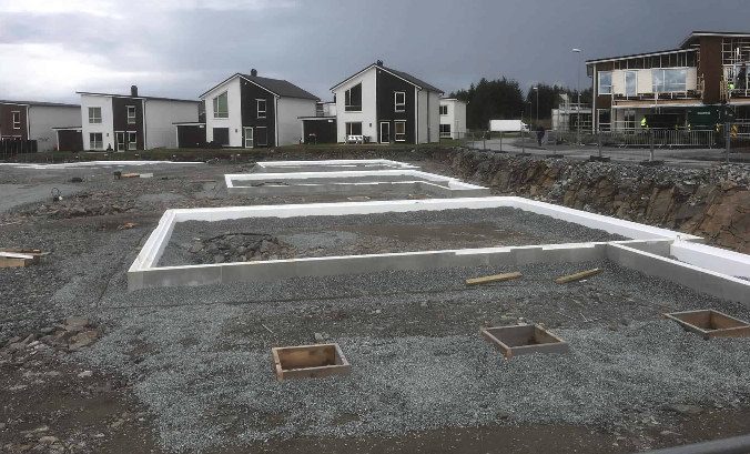 Byggeplass med grunnmur og flere hus i bakgrunnen under en mørk himmel.