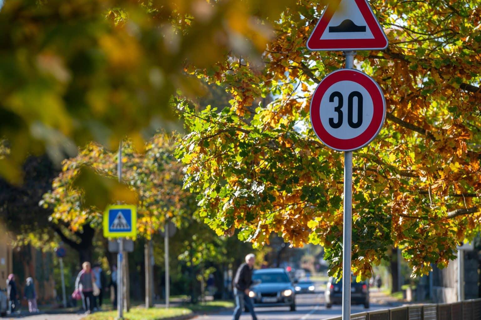 Skilt med hastighetsgrense på 30 km/t omgitt av høstfargede trær og fotgjengere på en gate.