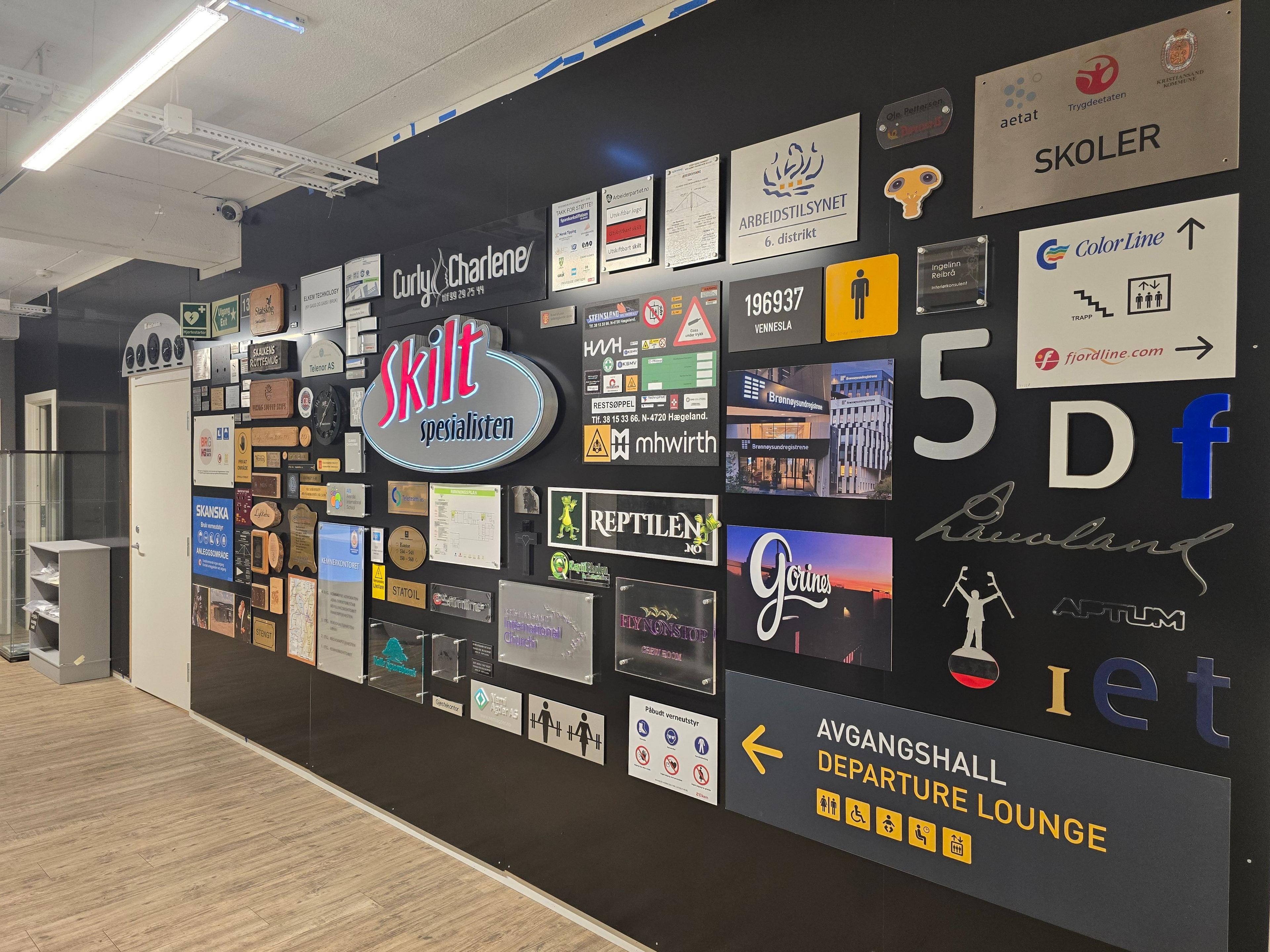 En vegg med ulike skilt og informasjonstavler i et moderne senter, inkludert avgangshall og butikker.