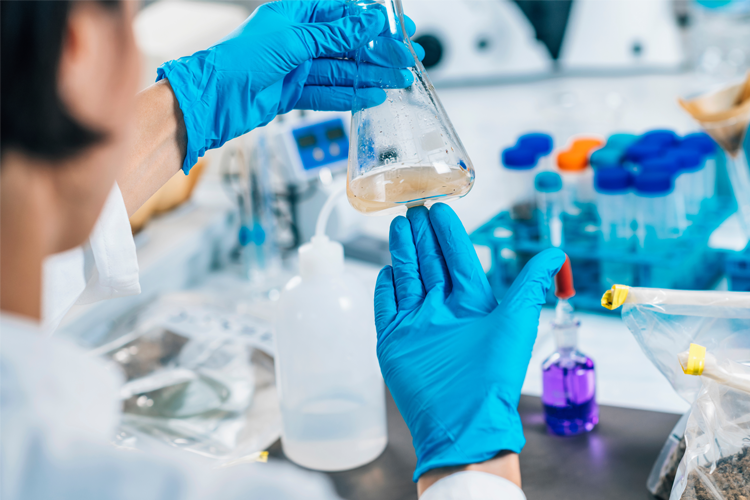 En person med blå hansker holder en kolbe med væske i et laboratorium, omgitt av laboratorieutstyr.