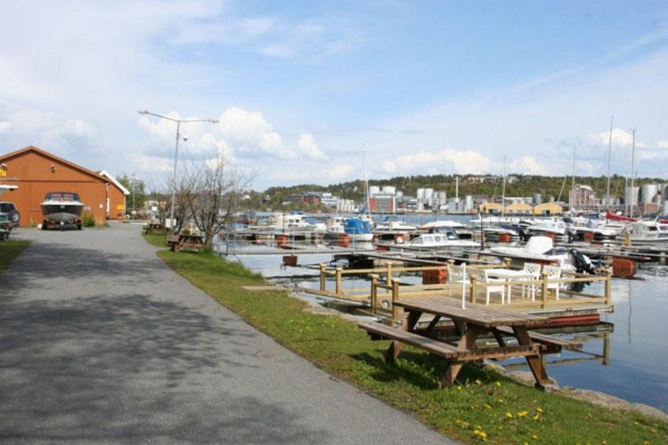 Bilde av en havn med båter, en gangvei og en rød bygning i bakgrunnen, under en blå himmel.