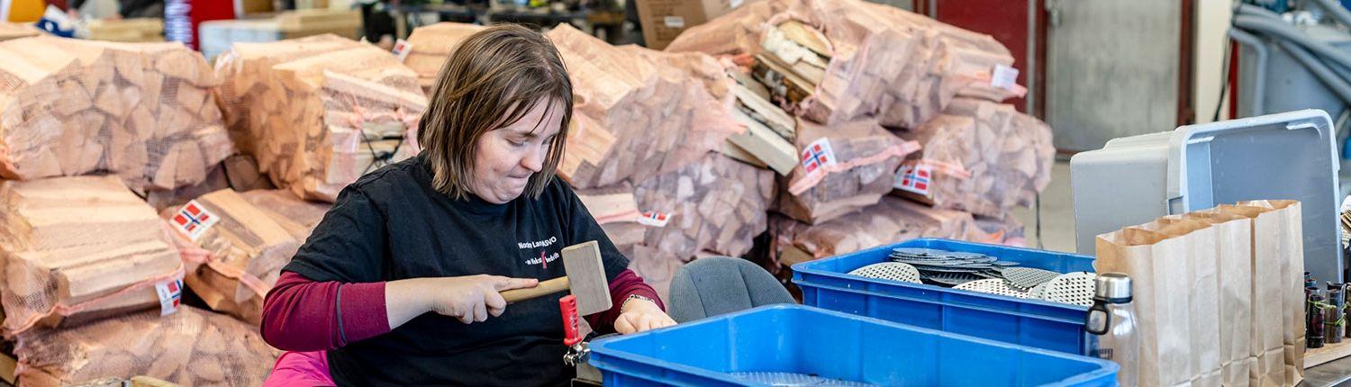 Kvinne som jobber med trebearbeiding i en fabrikk, omringet av treplanker og verktøy.