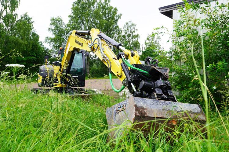 Gravemaskin i hagen med høyt gress og trær i bakgrunnen, klar til å utføre graving.