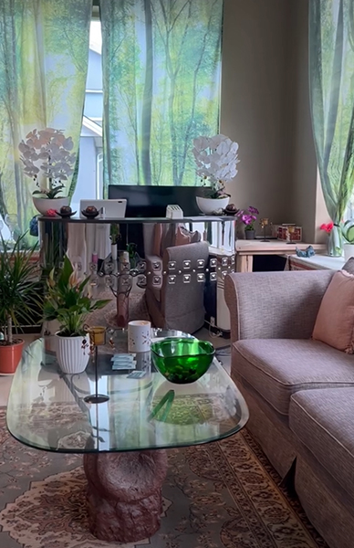 Moderne stue med glassbord, sofa, planter og gardiner med skogmotiv.