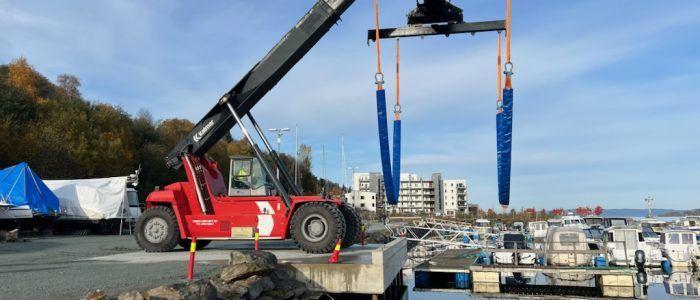 En rød kran løfter blå heiser ved en marina med båter og bygninger i bakgrunnen.