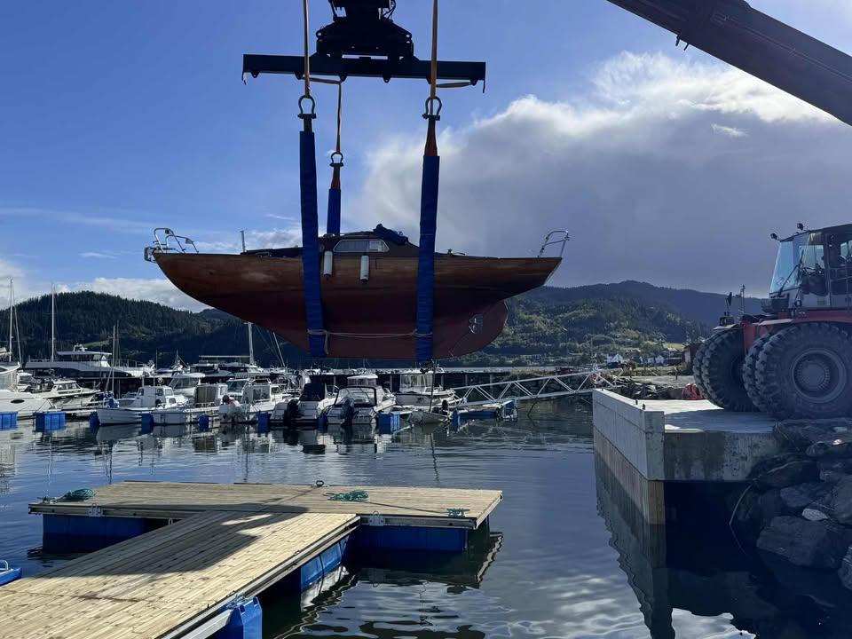 Båt heises opp av en kran ved en marina, med flere båter og fjell i bakgrunnen.