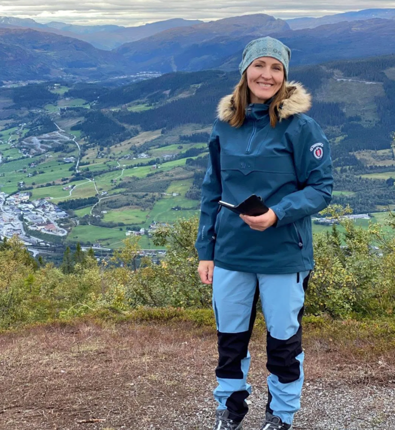 Kvinne i turklær står på en fjelltopp med vakker utsikt over dalen og landsbyen nedenfor.
