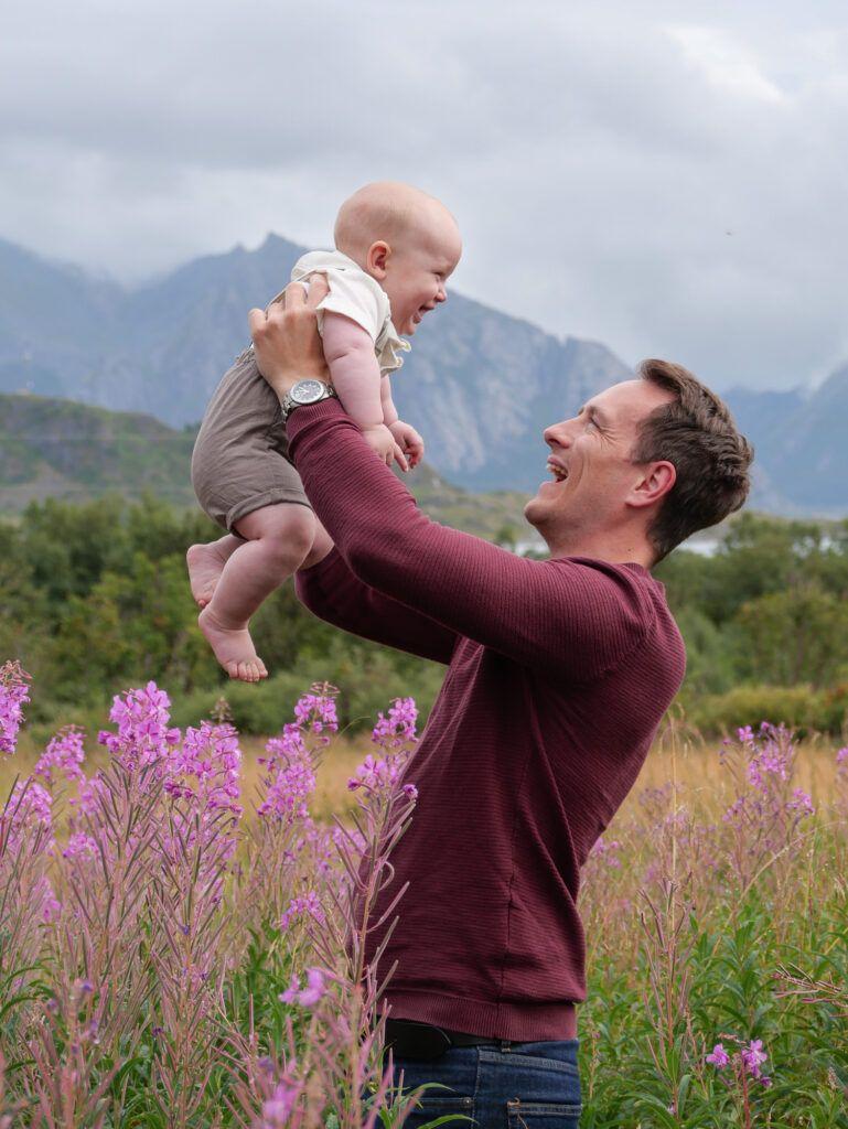 En far løfter sin baby opp i luften mens de står i en blomstereng med fjell i bakgrunnen.