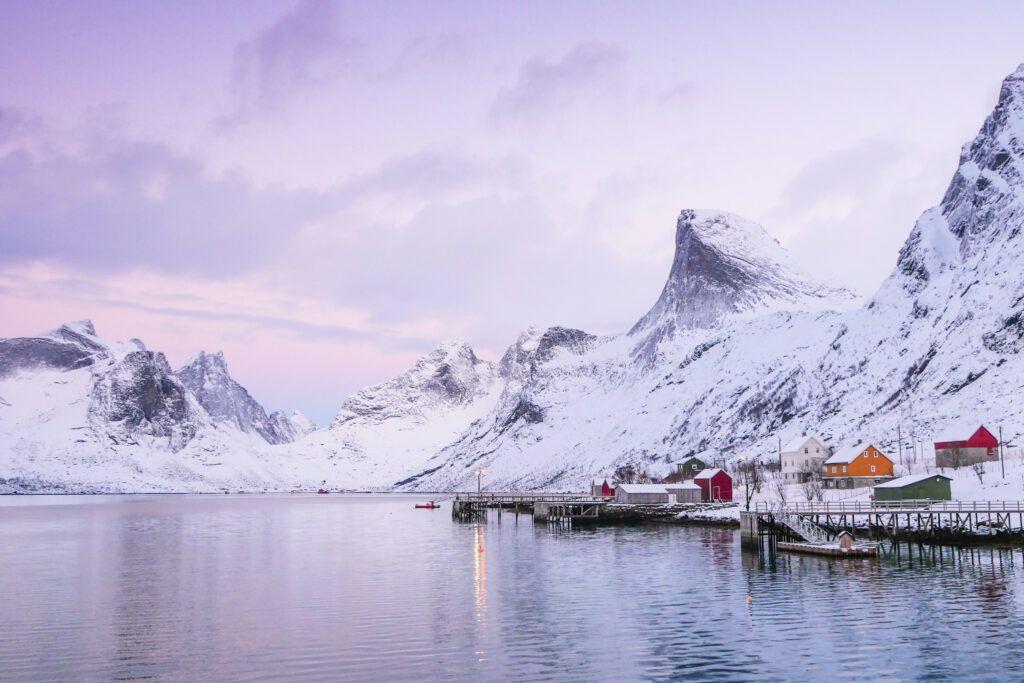 Vinterlandskap med snødekte fjell og fargerike hytter ved en rolig fjord i Norge.