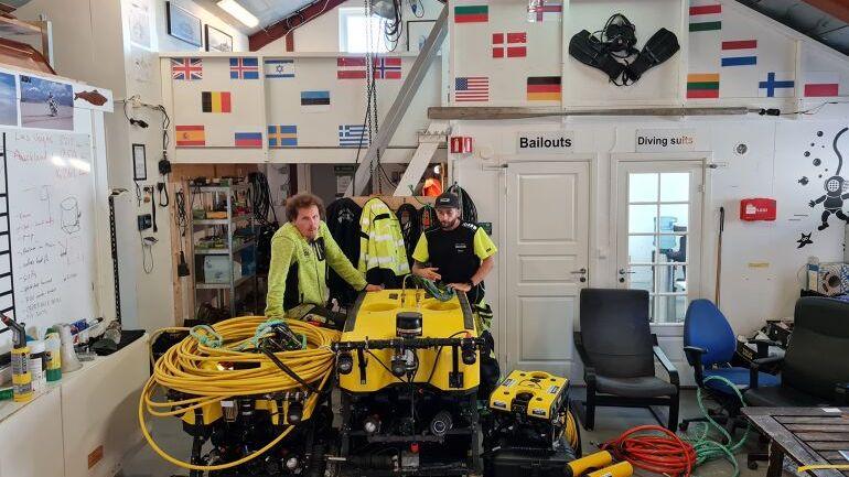 To personer i gule drakter står ved en undervannsdronning med kabler i et rom dekorert med flagg.
