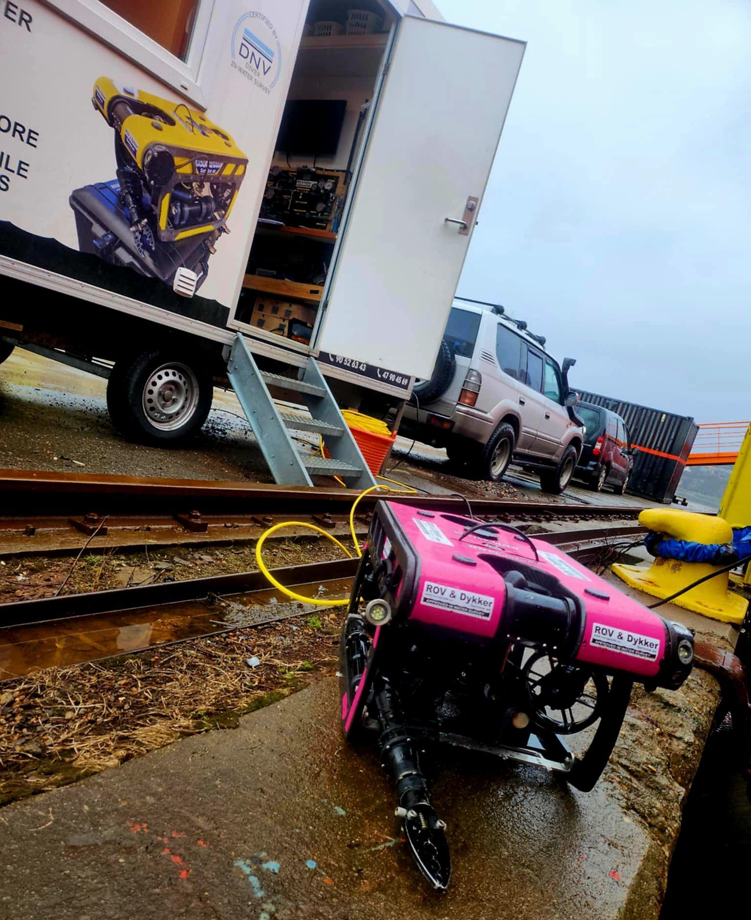 En rosa robot med slanger står på bakken ved en lastebil og en tilhenger på en byggeplass.