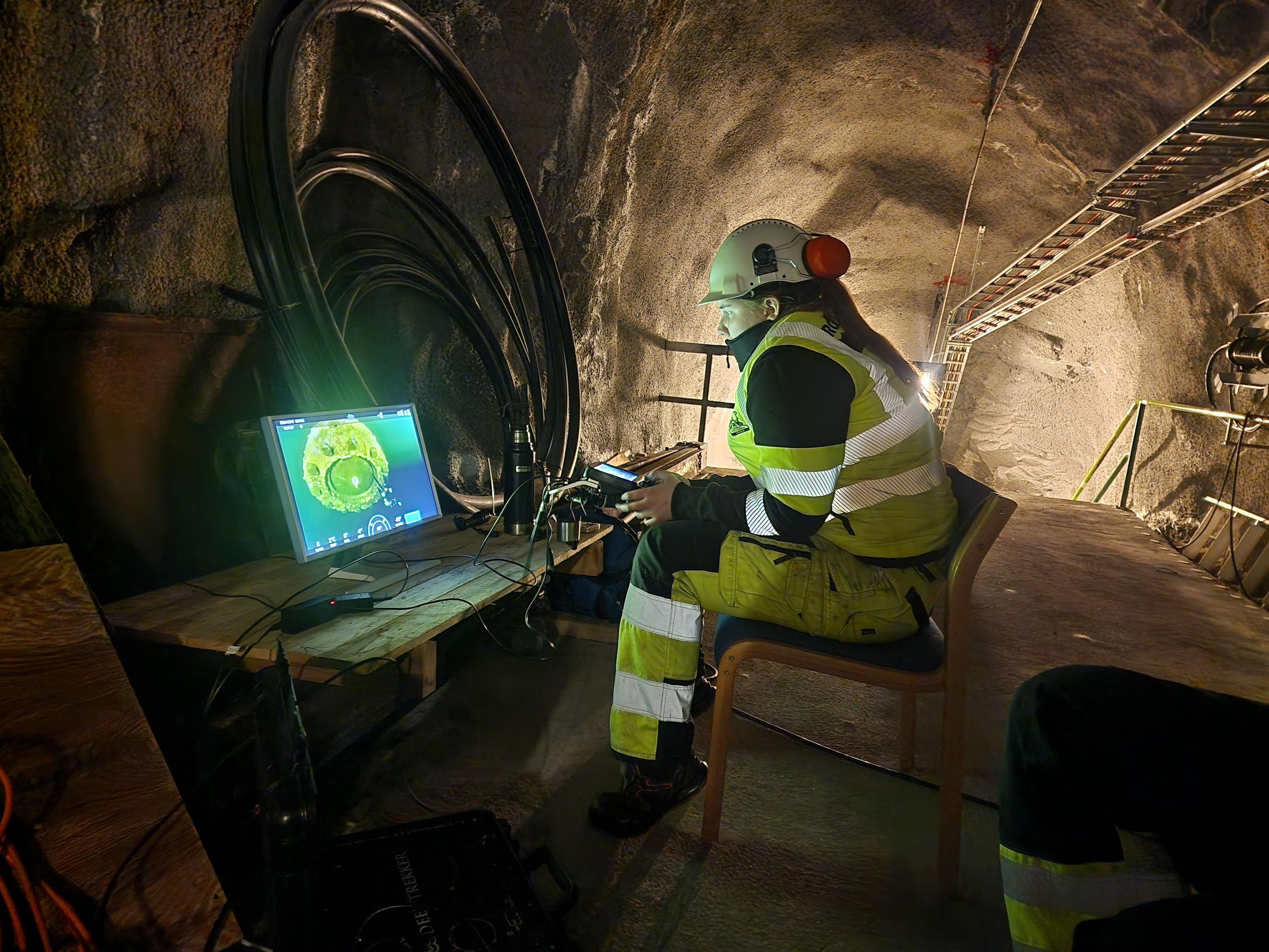 En arbeider i sikkerhetsutstyr sitter foran en datamaskin i en tunnel med kabler rundt seg.