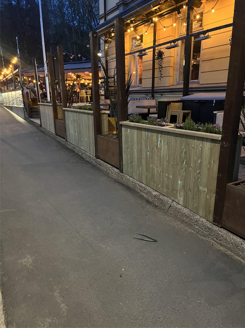 Uteområde med trepanel og belysning ved en restaurant, med sitteplasser og blomsterkasser langs fortauet.