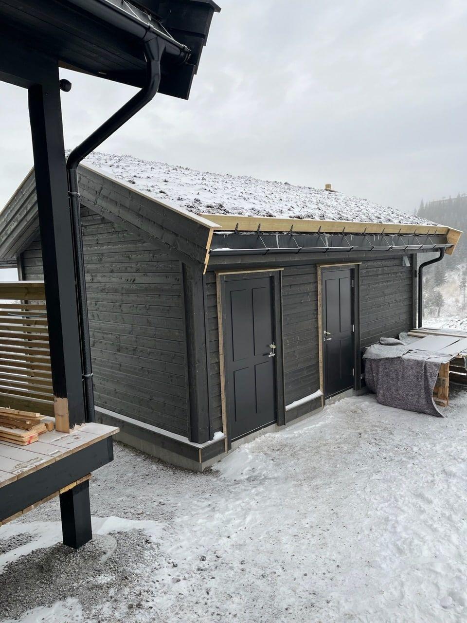 En moderne hytte med grå vegger og to dører, dekket av snø, omgitt av vinterlandskap.