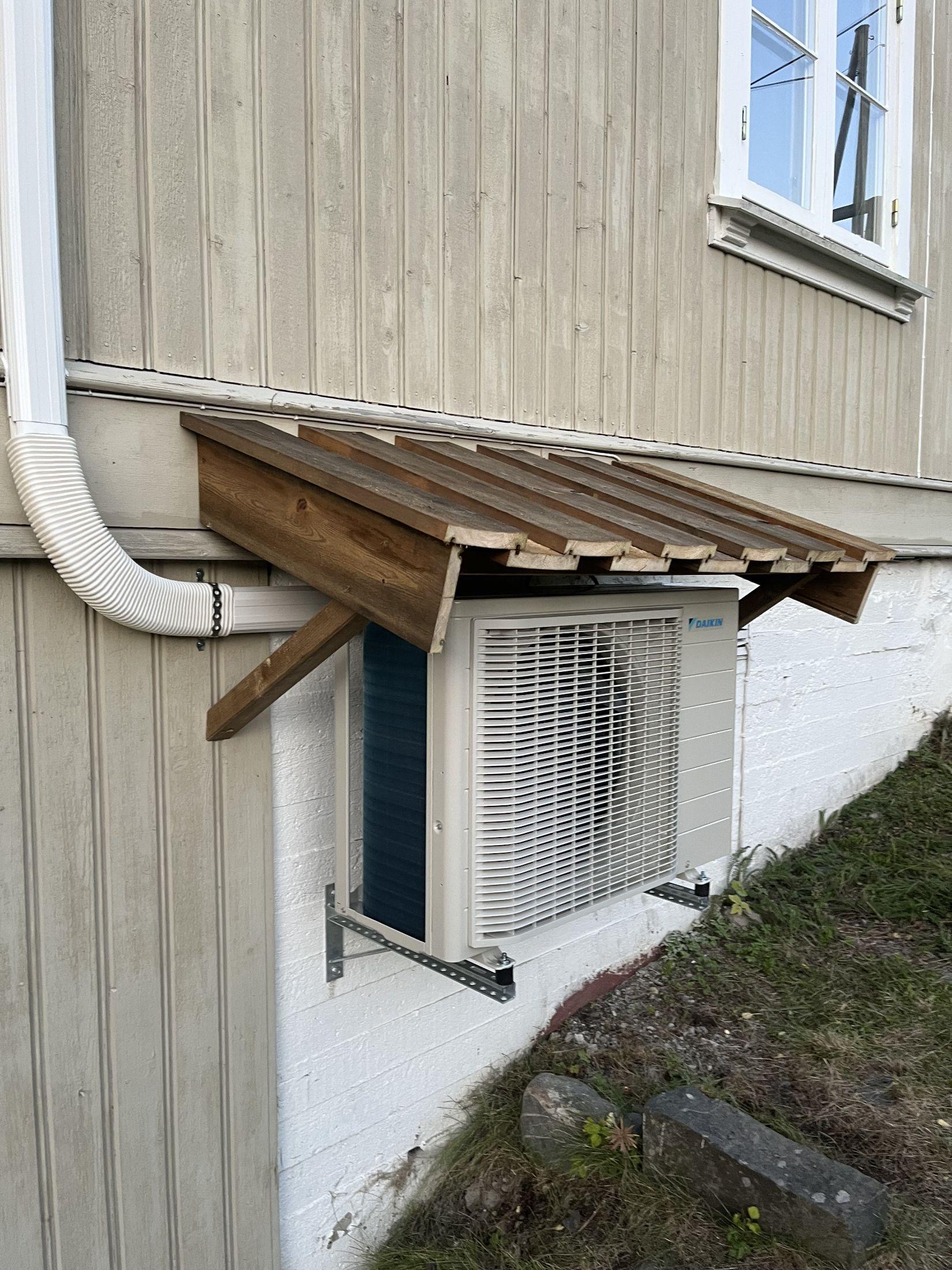 Klimaanlegg installert på veggen av et hus, beskyttet av et treoverbygg.