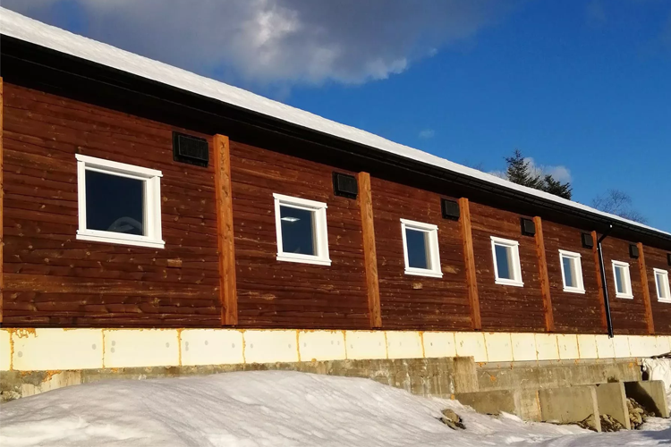 En moderne hytte med trepanel og hvite vinduer, delvis dekket av snø, under en blå himmel.